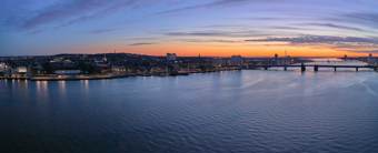 Aerial view of a city waterfront at sunset, with buildings, bridges, and calm water reflecting the evening sky.
