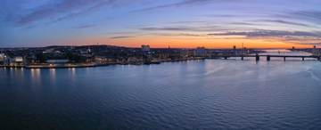 Aerial view of a city waterfront at sunset, with buildings, bridges, and calm water reflecting the evening sky.