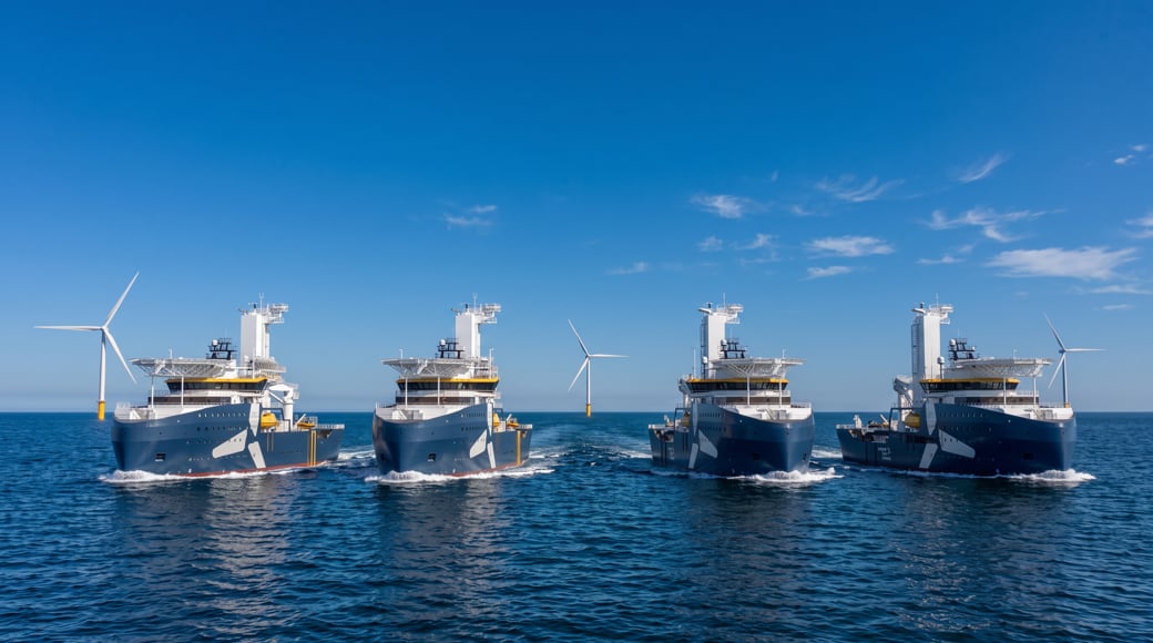 Four modern offshore vessels sailing side by side on a calm blue sea, with offshore wind turbines visible in the background under a clear sky.