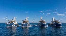 Four modern offshore vessels sailing side by side on a calm blue sea, with offshore wind turbines visible in the background under a clear sky.