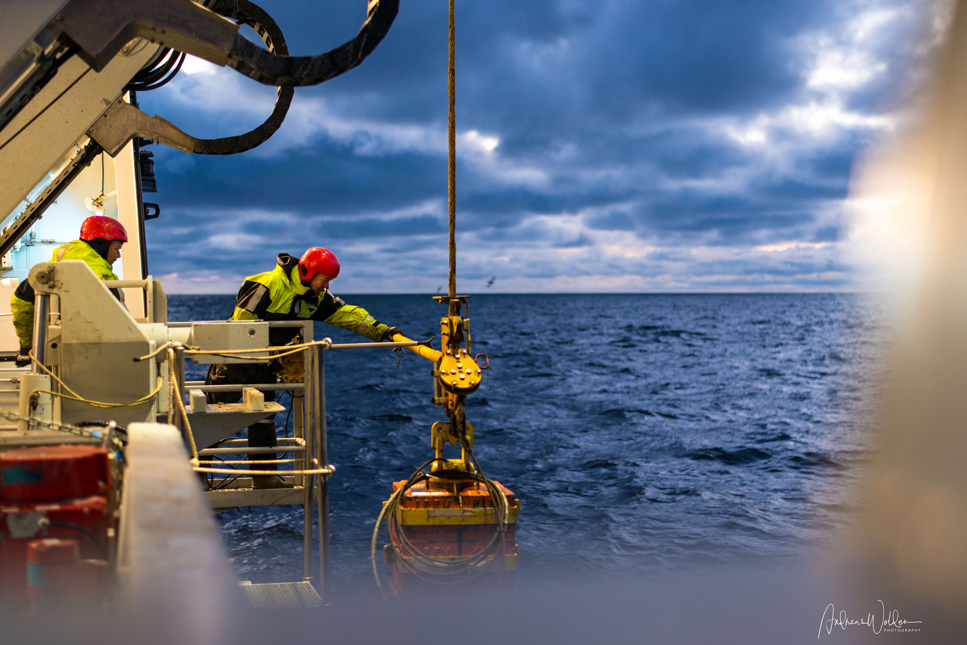 Two people handling an underwater robot that is hanging in a wire of the side of the vessel.
