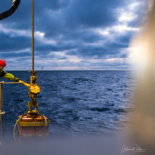 Two people handling an underwater robot that is hanging in a wire of the side of the vessel.