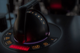Close-up of two joystick-style control levers mounted on a dark console, each surrounded by illuminated circular indicators in red and purple tones.