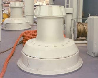 A large cylindrical marine deck winch is mounted on a ship’s deck, with a thick orange rope coiled beside it and a cable spool visible in the background. 