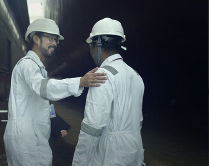 Two Kongsberg Maritime field service engineers dressed in white and with protective gear in front of a vessel hull in dry dock