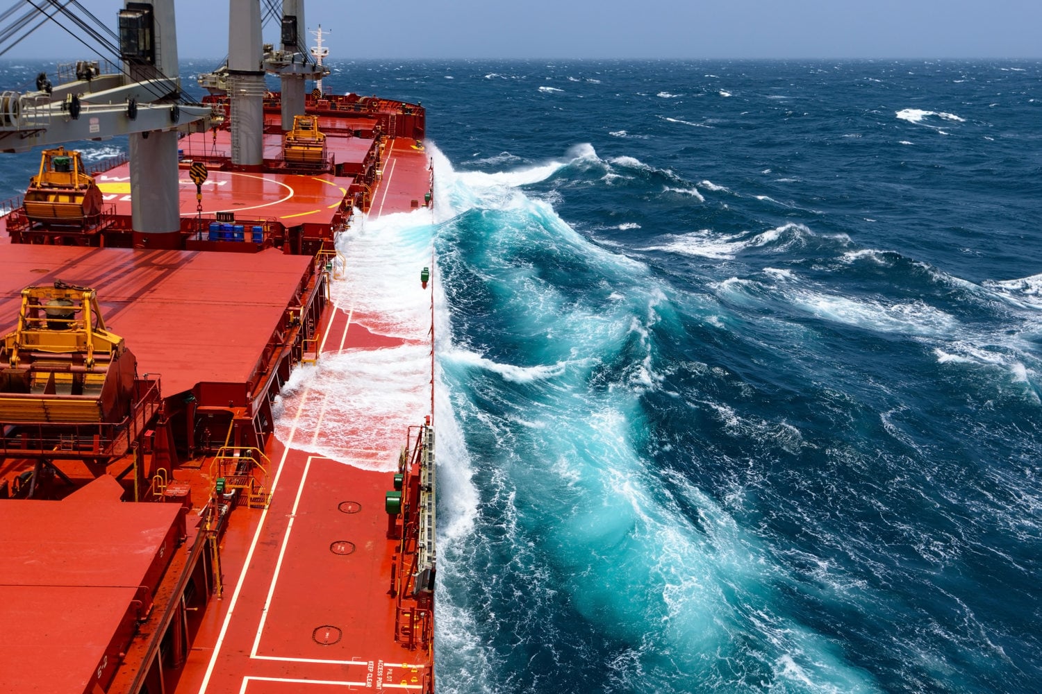 Large cargo vessel at sea in rough weather, with waves breaking along the red deck and white spray sweeping across the ship’s side