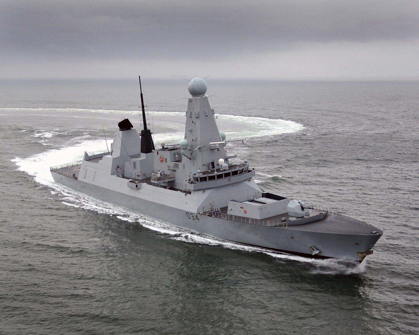 A grey naval vessel making a sharp turn at sea, leaving a circular wake pattern behind under an overcast sky.