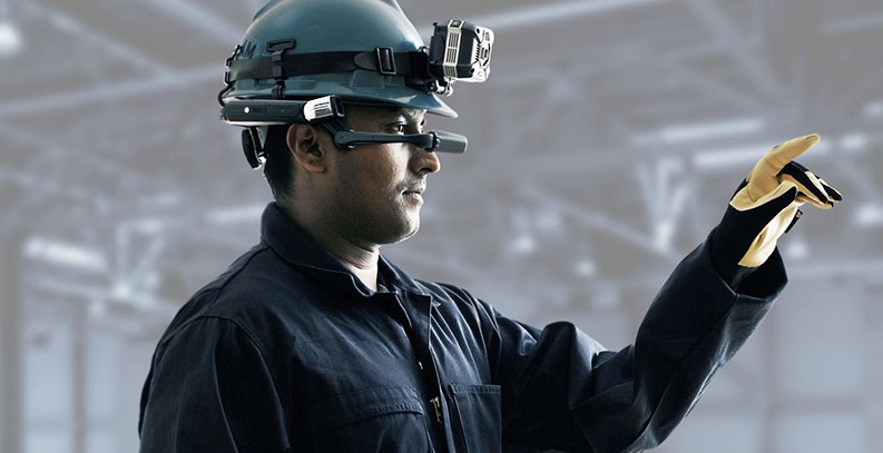 A vessel technician wearing protective gear, a headlight, and a mixed reality headset and pointing to equipment onboard to enable remote inspection and troubleshooting by a service expert