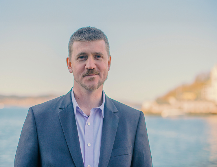 Person wearing a suit jacket and shirt, standing outdoors near water with a coastal landscape visible in the background.