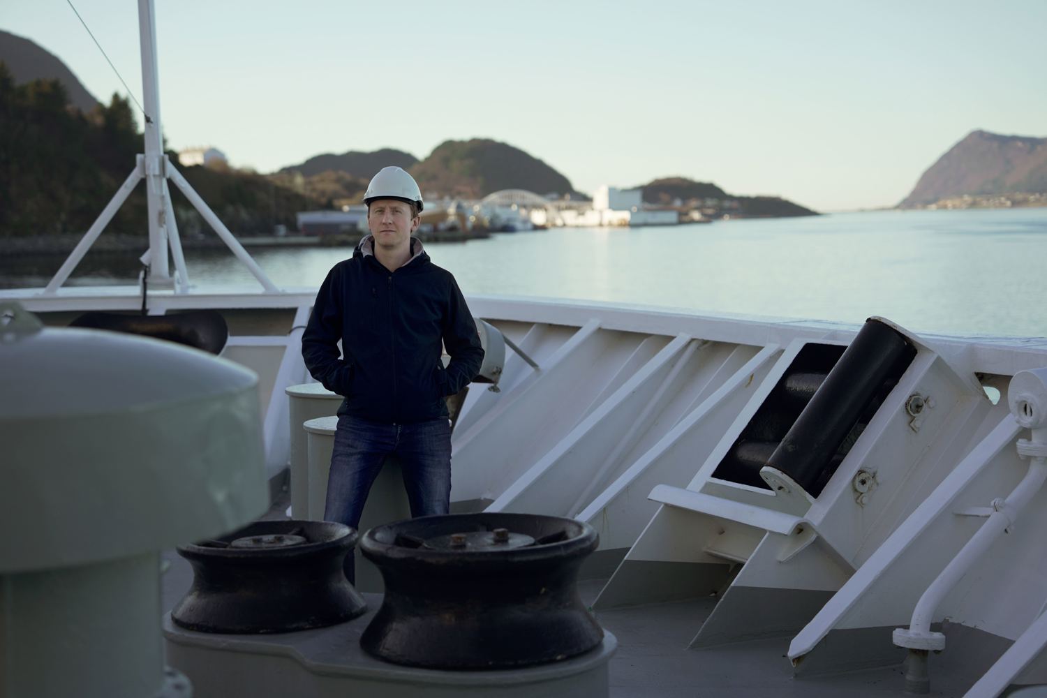 Employee wearing a hard hat standing on the deck of a vessel, with the coastline in the background.