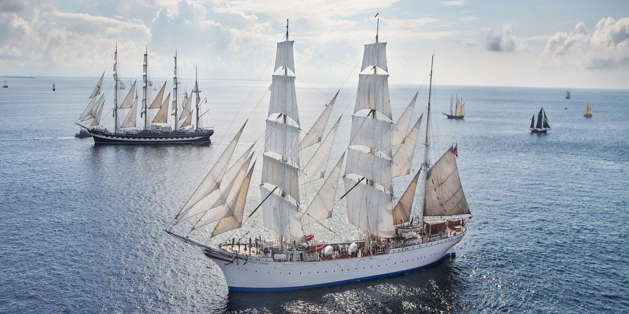 A birdseye view of Statsraad Lemkuhl in open ocean swith several other tall ships and sailboats in the background.