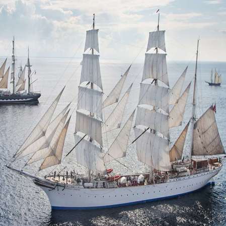 A birdseye view of Statsraad Lemkuhl in open ocean swith several other tall ships and sailboats in the background.