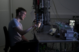 Person sitting at a workbench in a lab, interacting with a tablet connected to electronic equipment and a dense setup of wires, circuit boards, and testing devices on the desk behind.