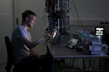 Person sitting at a workbench in a lab, interacting with a tablet connected to electronic equipment and a dense setup of wires, circuit boards, and testing devices on the desk behind.