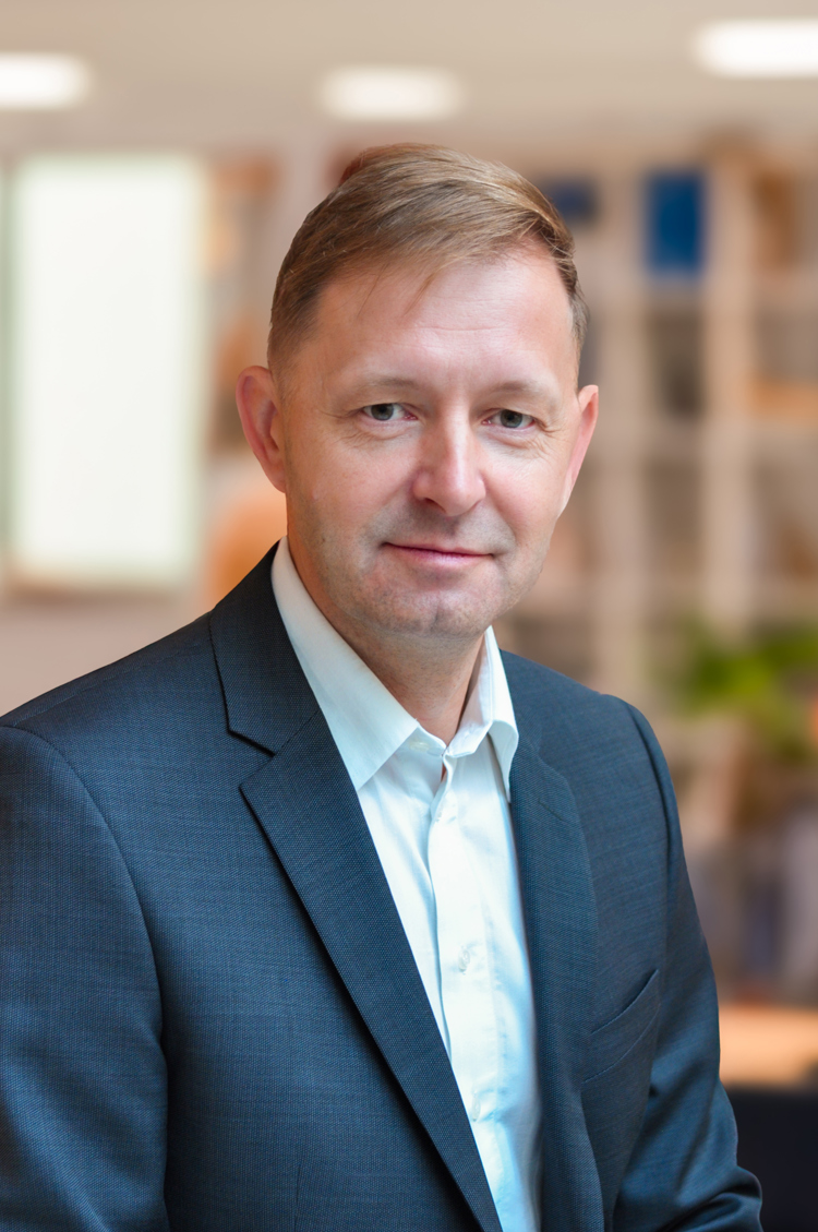 Upper‑body portrait of a person wearing a suit jacket and shirt, photographed indoors with a softly lit background.