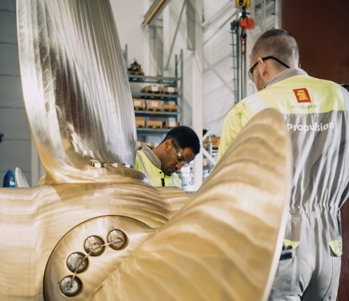 Two technicians in high-visibility workwear inspecting a massive bronze marine propeller inside an industrial workshop.