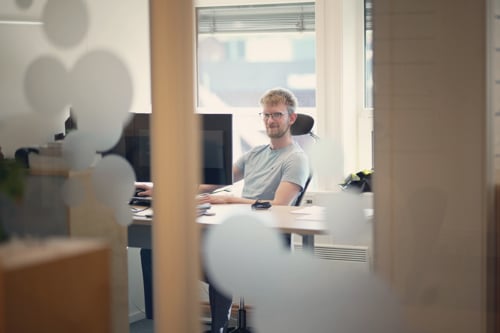 A picture outside a mans office where you can see him sitting on his desk in a blue t-shirt