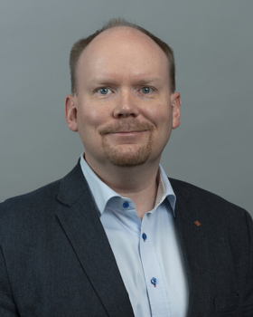 Portrait photograph of a person wearing a dark blazer and light blue shirt, shown against a plain grey background, cropped at the shoulders.