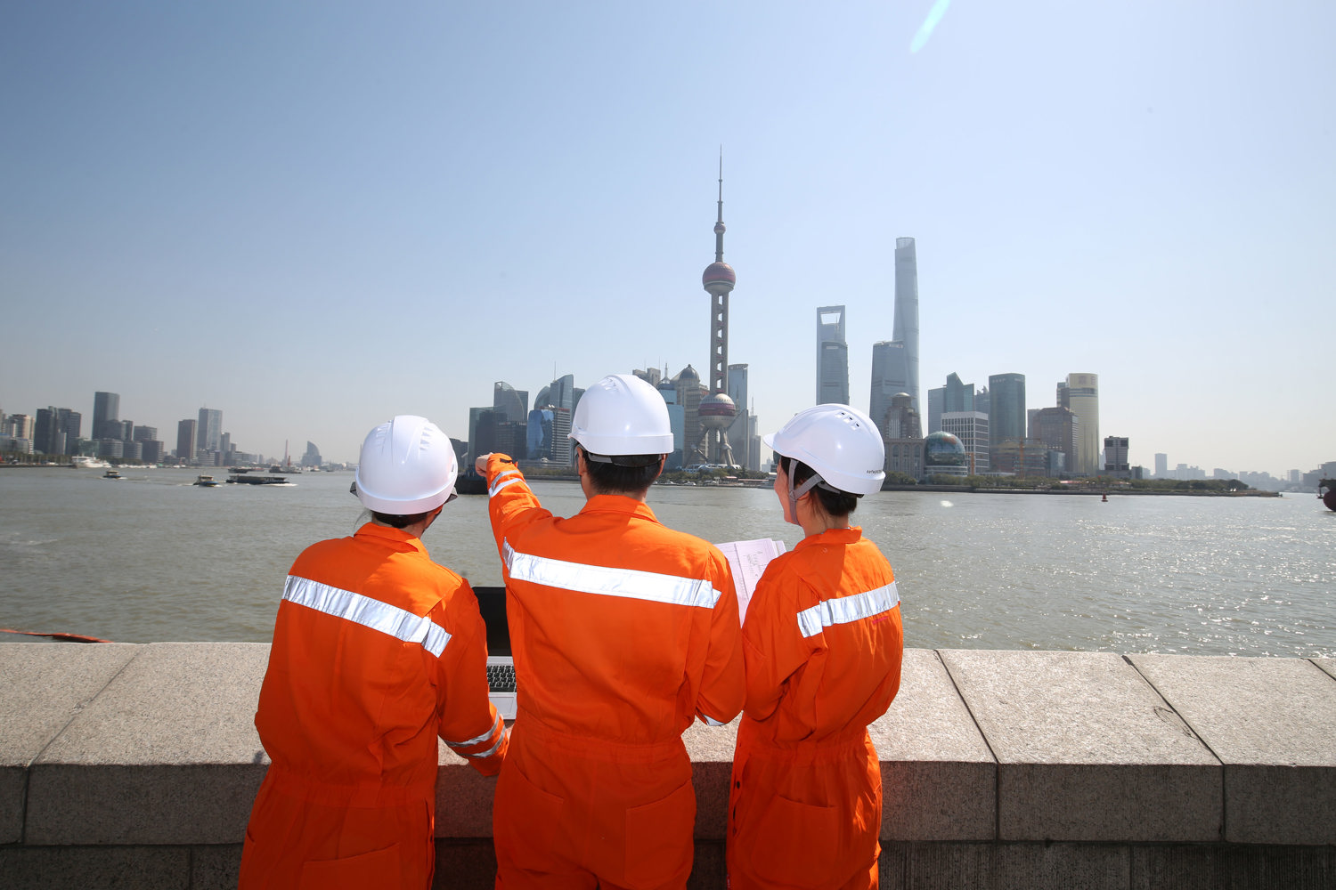 Three field service engineers standing in front of a maritime passage in an urban environment, pointing at vessels visible in the background