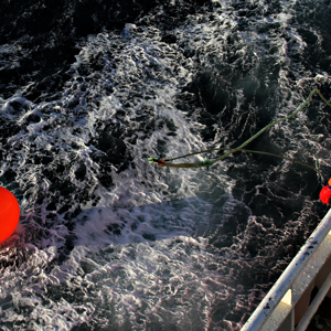 Fisherman in orange clothing standing on a ship looking at a buoy floating in the sea.