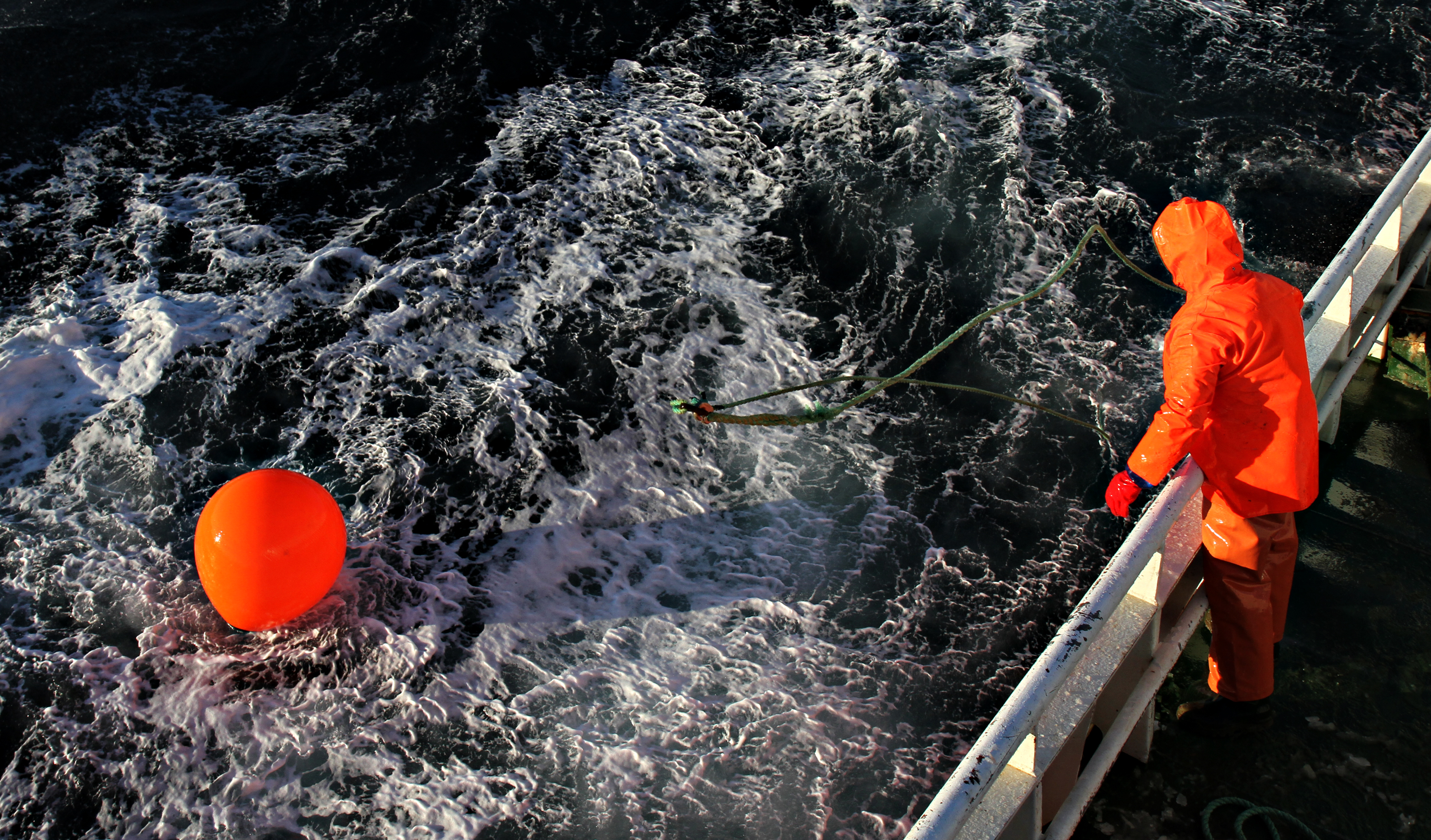Fisherman in orange clothing standing on a ship looking at a buoy floating in the sea.