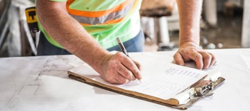 Close-up of a worker in a high-visibility vest writing notes on a clipboard placed over technical drawings in an industrial workshop