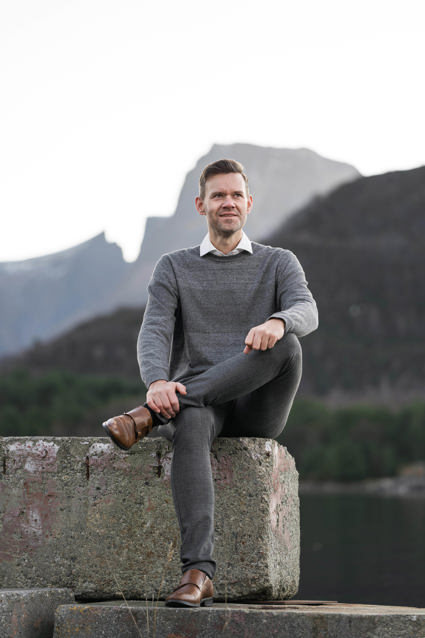 Person seated outdoors with legs crossed, wearing a sweater and collared shirt, with mountains and forested landscape in the background.