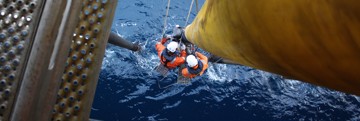 Technicians in protective equipment performing maintenance work on a large offshore riser or pipeline structure, suspended above the sea alongside an offshore installation